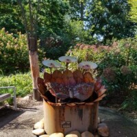 “Large rust-colored metal sculpture resembling a flower with petal-shaped elements and disc-like forms rising from the center at the Botanical Garden of the Ozarks, mounted on a cylindrical base surrounded by smooth stones, with trees, shrubs, and a bench in the background