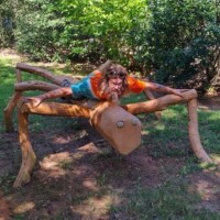 Large wooden spider sculpture with eight carved legs and circular eyes at the Botanical Garden of the Ozarks, with a person in a tie-dye shirt and sunglasses lying face down on top, arms extended along the front legs in a setup moment tagged for zach-chaos-duo-1