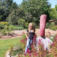 Oversized red-handled garden trowel sculpture embedded in the ground at the Botanical Garden of the Ozarks, with a person in sunglasses, black shirt, and jeans posing beside it in a playful moment, surrounded by lush plants and trees