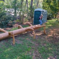 Large wooden log sculpture with protruding limbs placed horizontally on the ground at the Botanical Garden of the Ozarks, with a person balancing on top in a chaotic reaction moment tagged for zach-chaos-duo-2, portable toilets lined up in the background
