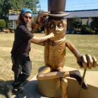 Mr Peanut statue Fort Smith Arkansas, golden sculpture of the iconic character seated with one leg crossed, wearing a top hat, monocle, gloves, and holding a cane, located outdoors near a building with a blue awning and surrounded by grass and bushes, with Zach playfully adjusting the statue’s hat and touching its face