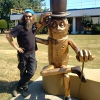 Mr Peanut statue Fort Smith Arkansas, seated sculpture of the iconic character with top hat, monocle, gloves, and cane, posed with one leg crossed and one arm resting on its knee, located outdoors near a building with large windows, surrounded by grass and trees, with Zach standing beside the statue wearing a black shirt and pants, one hand on the hat and the other on his hip