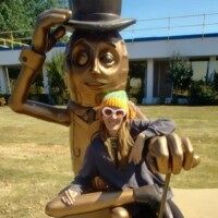 Mr Peanut statue Fort Smith Arkansas, bronze sculpture of the iconic character with top hat, monocle, gloves, and cane, posed outdoors near a building with large windows and surrounded by greenery, with Nancy leaning against the statue’s hand wearing a colorful knit hat and white-rimmed sunglasses, smiling