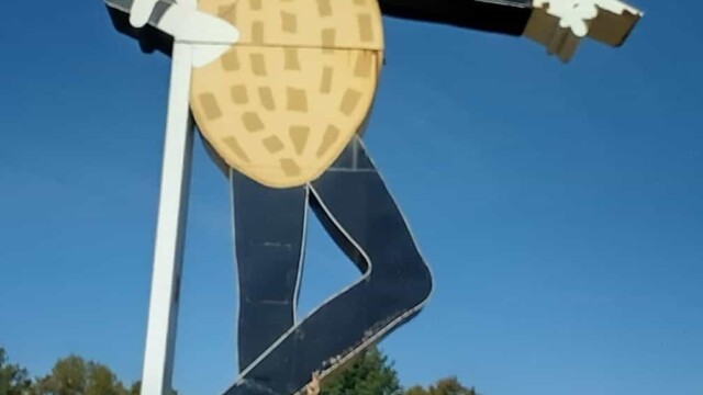 Mr Peanut statue Fort Smith Arkansas, featuring the iconic character in a top hat labeled ‘MR. PEANUT,’ white gloves, black pants, and white shoes, holding a cane and pointing, standing on a brick base in a grassy roadside area with trees and a road in the background