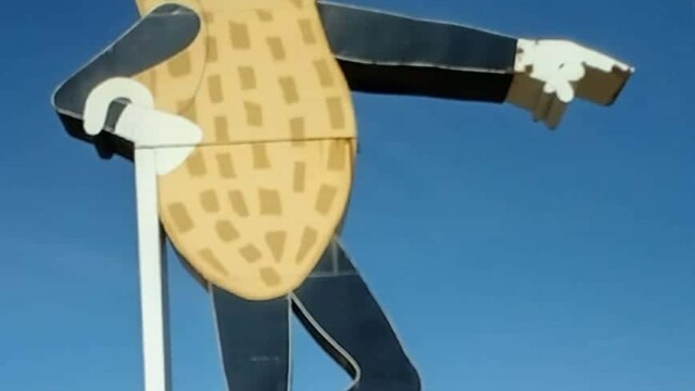 Mr Peanut statue Fort Smith Arkansas, showing the iconic character with monocle, top hat labeled ‘MR. PEANUT,’ gloves, and shoes, posed with one leg crossed and holding a cane, standing on a brick base in a grassy area with trees and buildings in the background