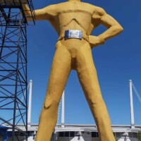 A person sitting on the left foot of the towering Golden Driller statue in Tulsa, Oklahoma, emphasizing its massive scale against the human-sized visitor.
