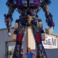 Optimus Prime statue at G&M Body Shop east location in Stillwater, Oklahoma, with a person posing in front — one arm raised, the other on hip. Painted in red, blue, and silver, the towering robot stands beside signage reading “A&M EAST.” Part of the Transformers statues Stillwater Oklahoma roadside display.