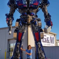 Optimus Prime statue at G&M Body Shop in Stillwater, Oklahoma, with a person posing in front, arm extended toward the towering robot. Constructed from metal parts and labeled “G&M GIANT” in the background, this figure anchors the east side of the Transformers statues Stillwater Oklahoma display.