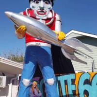 Giant cowboy statue holding a silver fish in Meadow Gold District Tulsa, with colorful Route 66 mural behind and a person standing at the base for scale under clear blue skies.