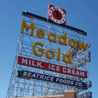 Meadow Gold District Tulsa landmark sign featuring yellow “Meadow Gold” neon letters, red “Milk • Ice Cream” panel, blue “Beatrice Foods Co.” panel, and a red clock mounted on a metal framework above a brick building under a clear blue sky.