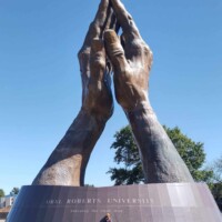 Massive bronze sculpture of two hands pressed together in prayer, mounted on a circular pedestal inscribed with “ORAL ROBERTS UNIVERSITY” and “PRAYING HANDS SCULPTURE.” A person stands at the base, emphasizing the scale. Clear blue sky and trees frame the outdoor setting.