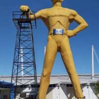 The Golden Driller statue in Tulsa, Oklahoma, standing tall with one hand resting on an oil derrick, against a clear blue sky and industrial backdrop.