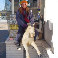 Person sitting on a white mechanical horse ride outside a storefront, wearing an orange beanie, white sunglasses, and oversized jacket, with a guitar and “Fear Farm” poster visible through the window.