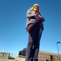 Person in a yellow hat, blue jacket, purple gloves, and dark pants standing with arms crossed on a paved area in downtown Tulsa, photographed from a low angle against a clear blue sky, with buildings and a streetlamp in the background — casual travel moment near the Center of the Universe Tulsa.