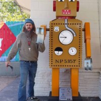 Person posing with a large cartoon-style robot sculpture featuring red ears, a chest gauge, and blocky limbs, set against a painted wall with geometric designs and a white brick backdrop.