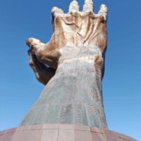 Large sculpted hand emerging from a dome-like tiled base, fingers extended upward against a clear blue sky. Realistic texture and scale dominate the frame, with no visible signage or context markers. Dramatic gesture and clean silhouette make it visually distinct from the world's largest praying hands installation.
