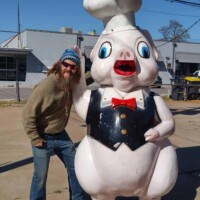 Visitor posing with a chef pig statue in Meadow Gold District Tulsa, featuring a black vest, red bow tie, and white hat, with street buildings and power lines in the background