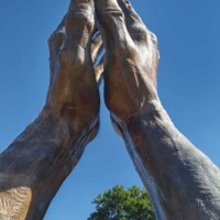 Close-up of the world's largest praying hands sculpture at Oral Roberts University, with bronze hands pressed together in prayer against a clear blue sky. Partial inscription visible at the base, reading “ERTS UNIVER,” hinting at the full campus name. Realistic texture and scale dominate the frame.