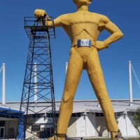 The Golden Driller, towering in his signature yellow outfit and hard hat, stands beside an oil derrick in Tulsa, Oklahoma, representing the city's petroleum industry legacy
