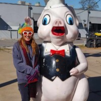 Person posing with a large pig statue dressed as a chef, featuring exaggerated facial features, a red bow tie, and a tall white hat, in a paved outdoor area with buildings and utility lines in the background.