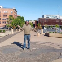Person in a cap, sunglasses, green long‑sleeve shirt, and jeans standing in the center of a circular brick and stone pattern in downtown Tulsa, arms slightly raised, surrounded by planters and multi‑story buildings under a clear blue sky — photographed at the Center of the Universe Tulsa.