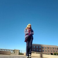 Person in a yellow hat, blue jacket, dark pants, and sneakers standing on a paved area in downtown Tulsa, with a multi‑story brick building, railing, and clear blue sky in the background — casual travel moment near the Center of the Universe Tulsa.
