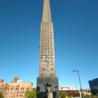 Triangular monument in downtown Tulsa with a flat disc top, inscribed vertically in multiple languages, with a person standing at its base; trees, buildings, and a clear blue sky in the background — part of the Center of the Universe Tulsa area.