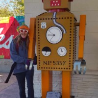 Meadow Gold District Tulsa sculpture of a cartoon-style robot with red ears, chest gauges, and blocky limbs, labeled “NP 5357,” with a visitor posing beside it against a painted wall and brick backdrop.