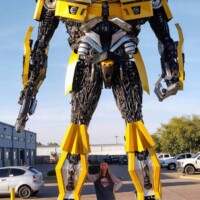 Bumblebee statue at G&M Body Shop west location in Stillwater, Oklahoma, with a person standing directly in front for scale. Painted in yellow with black and silver accents, the robot features gears, panels, and weapon-like arms. Cars, building, and blue sky frame the Transformers statues Stillwater Oklahoma roadside display