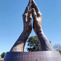 Wide-angle view of the world's largest praying hands sculpture at Oral Roberts University, with a person standing at the base for scale. Bronze hands pressed together in prayer, mounted on a circular pedestal inscribed with “EDUCATING THE WHOLE PERSON.” Clear sky and trees frame the outdoor setting.