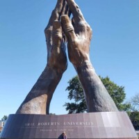 Outdoor view of the world's largest praying hands sculpture at Oral Roberts University, with bronze hands pressed together atop a circular base. Pedestal inscription reads “EDUCATING THE WHOLE MAN” and “SPIRIT – MIND – BODY.” A person stands at the base, highlighting the scale. Trees and blue sky frame the background.