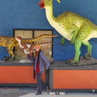 Person in colorful hat posing with three outdoor dinosaur statues, including a green and yellow figure with red mouth and two smaller orange and white models, set against a blue wall and orange platforms.