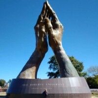 Wide shot of the world's largest praying hands sculpture at Oral Roberts University, with bronze hands clasped in prayer atop a grassy lawn. A person stands at the base for scale. Trees and a clear blue sky frame the outdoor setting. “ORAL ROBERTS UNIVERSITY” is inscribed on the pedestal.