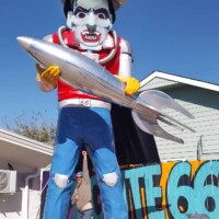 Oversized cowboy statue holding a silver fish and fork, with a person reaching up to touch its hand, set in front of a bold Route 66 mural on a sunny day with brick platform and building backdrop.