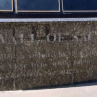 Dark stone monument labeled “WALL OF SACRIFICE,” engraved with a dedication to those who made the ultimate sacrifice in service to the country. Reflections obscure some of the text, but the inscription honors courage and valor. No visible figures or surrounding context.