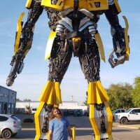 Bumblebee statue at G&M Body Shop west location in Stillwater, Oklahoma, towering over a person in blue shirt and jeans. Painted in yellow with black and silver accents, the robot stands on a paved lot with cars and buildings in the background. Part of the Transformers statues Stillwater Oklahoma roadside display.