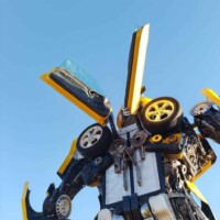 Bumblebee statue at G&M Body Shop west location in Stillwater, Oklahoma, constructed from yellow and black car parts including doors, wheels, and panels. Robot stands tall against a clear blue sky, with symmetrical automotive elements forming arms, legs, and head. Part of the Transformers statues Stillwater Oklahoma roadside display.