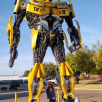 Bumblebee statue at G&M Body Shop west location in Stillwater, Oklahoma, with a person standing beneath it for scale. Painted in yellow with black accents, the robot features gears, pistons, and armor plating. Trees and building in background frame the Transformers statues Stillwater Oklahoma roadside display.