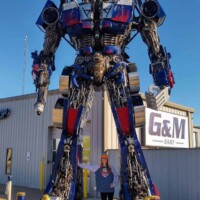 Full-body shot of the Optimus Prime statue at G&M Body Shop in Stillwater, Oklahoma, towering over a person with arms raised beneath it. Constructed from metal parts and painted in red, blue, and silver, this 22-foot-tall figure anchors the east side of the Transformers statues Stillwater Oklahoma display.