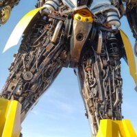 Low-angle view of the Bumblebee statue at G&M Body Shop west location in Stillwater, Oklahoma. Painted in yellow and black, the robot features gears, pipes, and metal panels arranged in a humanoid form. Clear blue sky and surrounding buildings frame the Transformers statues Stillwater Oklahoma roadside display.