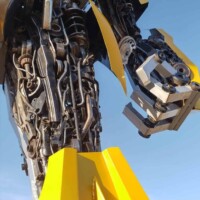 Low-angle close-up of the Bumblebee statue at G&M Body Shop west location in Stillwater, Oklahoma. Yellow and black metal panels, gears, and chains form the robot’s upper body, with clear blue sky in the background. Part of the Transformers statues Stillwater Oklahoma roadside display.