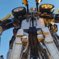 Bumblebee statue at G&M Body Shop west location in Stillwater, Oklahoma, viewed from a low angle against a clear blue sky. Yellow and black car doors extend upward like wings, with wheels and metal panels forming the robot’s chest and limbs. Part of the Transformers statues Stillwater Oklahoma roadside display.