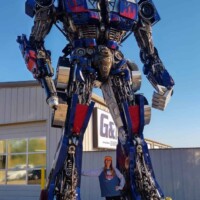 Optimus Prime statue at G&M Body Shop in Stillwater, Oklahoma, standing on a concrete pad with a person beneath it for scale. Painted in red, blue, and silver, the robot is flanked by two smaller mechanical figures near its feet. Partial signage reading “GA” visible in the background. Part of the Transformers statues Stillwater Oklahoma roadside display.