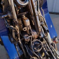 Close-up of mechanical components near the base of the Optimus Prime statue at G&M Body Shop in Stillwater, Oklahoma. Exposed gears, chains, and rods suggest motorcycle or vehicle parts repurposed into the larger Transformers statues Stillwater Oklahoma display. Blue-painted surface and corrugated metal wall in background.