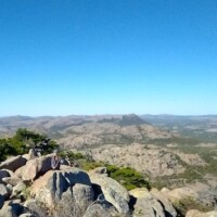 Panoramic view from Mount Scott’s summit in Oklahoma, with rugged granite boulders in the foreground, a lone hiker seated among them, and rolling Wichita Mountains stretching into the distance under a clear blue sky.