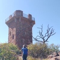 Person walking up a rocky trail toward a hexagonal stone watchtower with a flat roof and railing, framed by a twisted leafless tree against a clear blue sky.