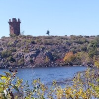 Brick watchtower with battlements perched on a rocky hill above a lake, framed by shrubs and a clear blue sky.