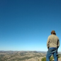 Person standing on a rocky outcrop at Mount Scott, Oklahoma, facing away toward a sweeping view of rugged hills and valleys under a bright blue sky.