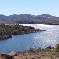 View across a shimmering lake toward the Jed Johnson Watchtower Lawton Oklahoma, with rocky hills and sparse vegetation under a bright blue sky.