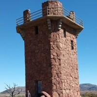 Jed Johnson Watchtower Lawton Oklahoma — cylindrical stone tower with crenellated top and metal railing, standing in the Wichita Mountains landscape, with a person at the base for scale.